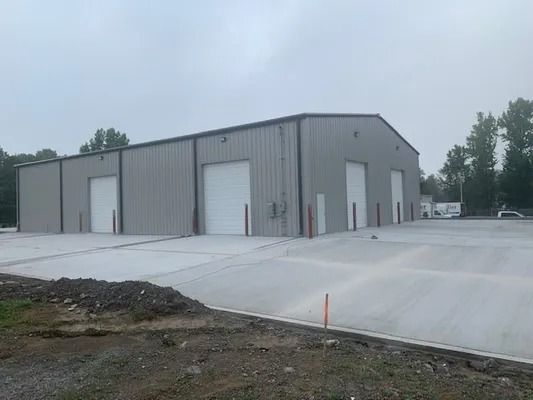 A grey metal warehouse with four closed roll-up doors, fronted by a large, newly poured concrete parking area.