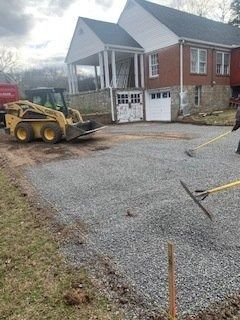 A construction worker uses a skid steer and rakes to level gravel for a driveway in front of a brick and stone house.
