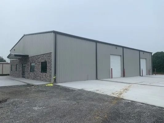 A gray, metal-sided commercial building with stone facade, two white roll-up doors, and a gravel parking lot.