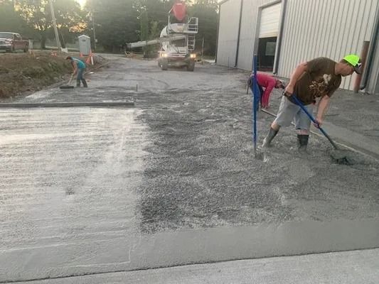 Workers use rakes and tools to smooth freshly poured concrete in an outdoor area beside a large building.