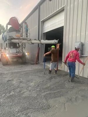 Three workers use tools to spread wet concrete poured from a truck chute in front of a metal industrial building.