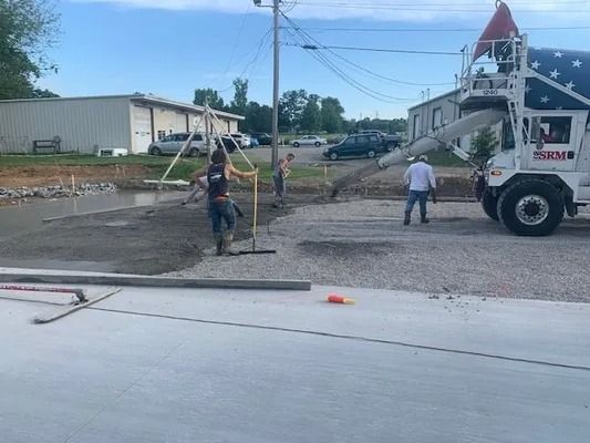 Construction workers smooth wet concrete being poured from a truck onto a gravel surface near a commercial building.