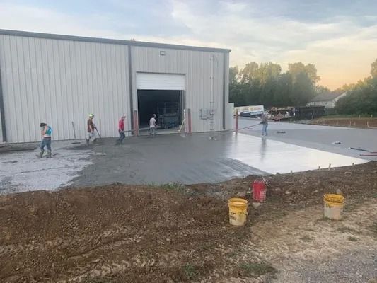A construction crew smooths freshly poured wet concrete in front of a large metal warehouse building at dusk.