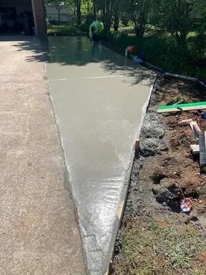 Freshly poured concrete forming a triangular extension along an existing driveway, with tools nearby on bare soil.