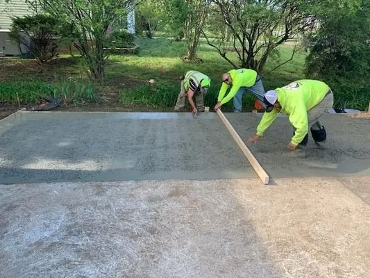 Three workers in neon green shirts use a long board to level freshly poured wet concrete outdoors.