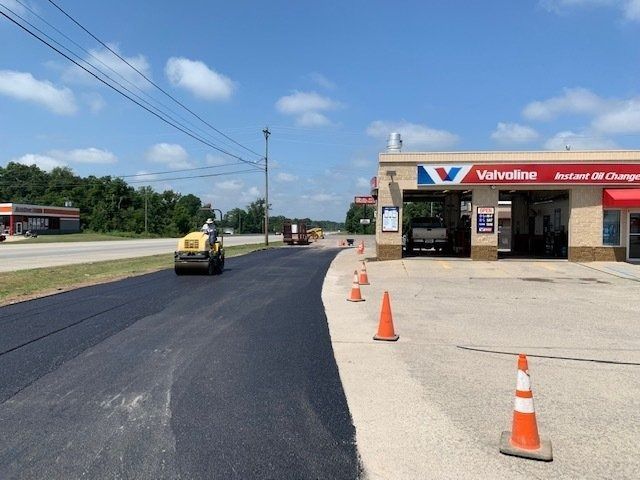 A construction roller compacts fresh asphalt on a driveway in front of a Valvoline Instant Oil Change station.