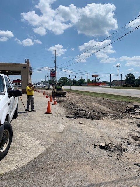 A person in a yellow shirt stands near orange cones at a road construction site with heavy equipment visible ahead.
