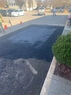 A rectangular section of newly laid dark asphalt sits in a parking lot, with a worker and parked cars visible in the back.