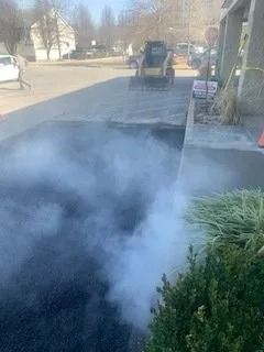 Thick steam rises from newly laid black asphalt in a driveway, with a small construction vehicle visible in the background.