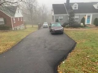 A freshly paved dark asphalt driveway leads toward a house with a parked dark-colored SUV on a foggy, overcast day.