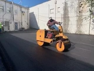 A person operates a yellow steamroller to smooth newly laid asphalt in an alley beside a white brick building.