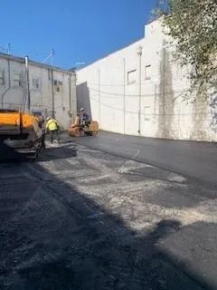 Workers operate a steamroller to pave an asphalt road between two light-colored buildings on a sunny day.