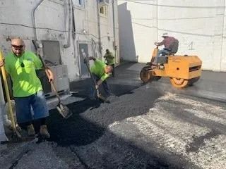Construction workers in bright green shirts pave a section of asphalt between two white buildings using a small roller.
