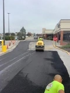 A construction worker in a lime-green shirt operates a yellow steamroller to pave a new asphalt section in a parking lot.