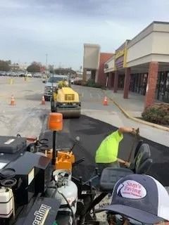 A construction worker in a neon shirt paves a parking lot with machinery, as a yellow steamroller approaches nearby.