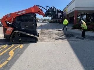 An orange Kubota skid-steer loader uses a grinder attachment to clear a patch of pavement near a building while workers stand by.