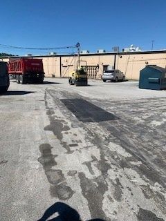 A yellow construction roller drives across an asphalt lot near a parked red dump truck, a car, and a blue dumpster.