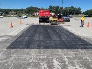 A construction crew repaving a section of a parking lot with a dump truck and a steamroller on a sunny day.