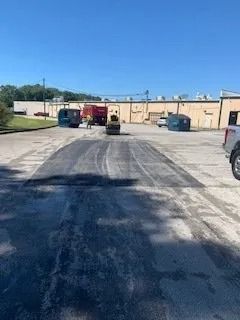 A construction roller compacts fresh black asphalt in a parking lot near an industrial building.