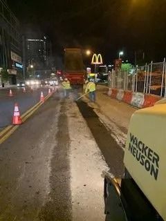 Road crew members paving a street at night, with a McDonald’s sign in the background and orange traffic cones nearby.