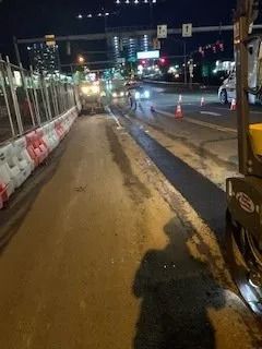 Night road construction site with freshly laid asphalt, barrier walls, and traffic cones illuminated by streetlights.