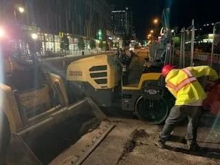 A night view of a construction worker in a high-visibility jacket using a yellow asphalt roller on a city street.