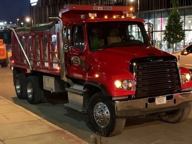 A red dump truck parked on a street corner during twilight with its headlights on.