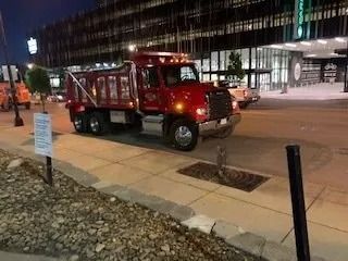 A red dump truck parked on a sidewalk in front of a modern building at dusk.