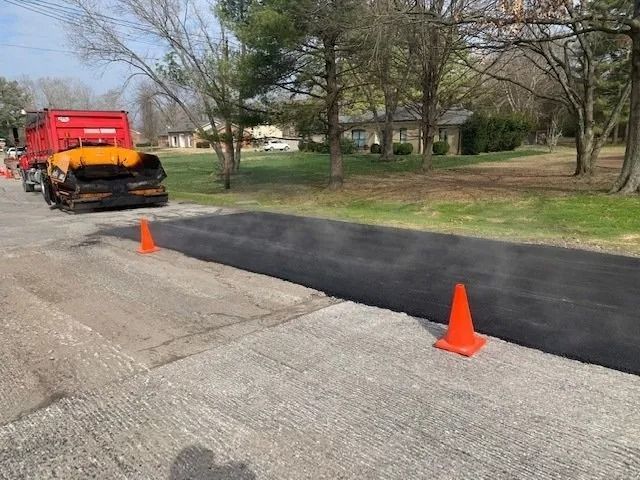 A red paving truck sits near a patch of fresh black asphalt on a street marked by two orange safety cones.
