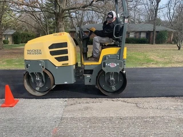 A worker operates a yellow and gray Wacker Neuson asphalt roller on a paved road next to an orange traffic cone.