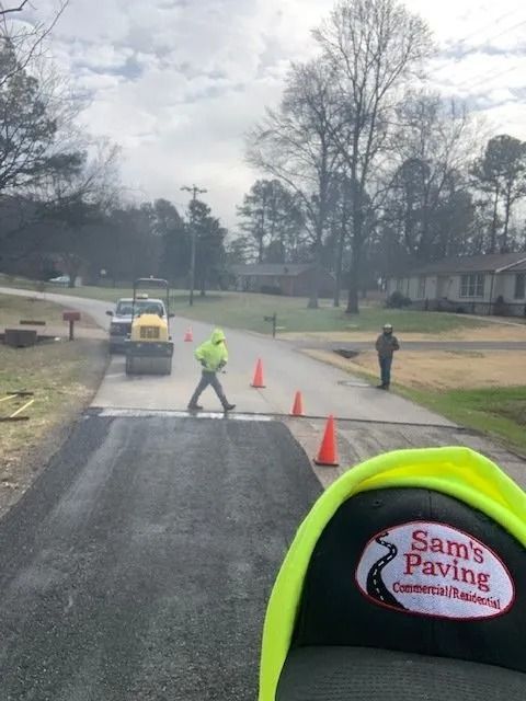 A crew member in a neon jacket works on a residential paving project, with a roller and traffic cones on a suburban street.