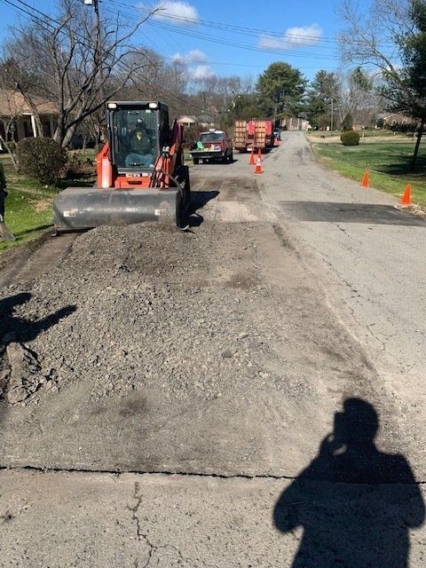 An orange skid steer loader sits on a residential street next to a patch of gravel and orange traffic cones.