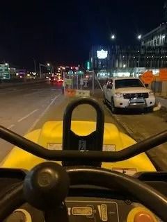 POV from inside a yellow construction vehicle at night, facing a parked white truck on a city street near road work.