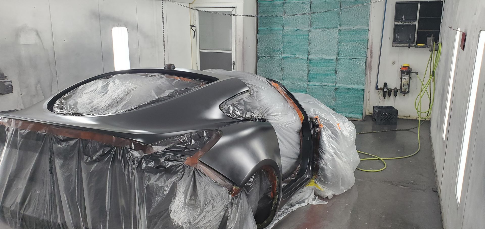 A gray sports car, covered in plastic, in a spray painting booth.