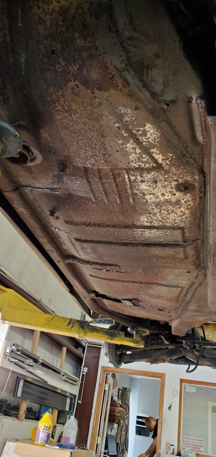 Underside of a car's rusty floor pan, viewed from below. The vehicle is on a lift, showing workshop setting.