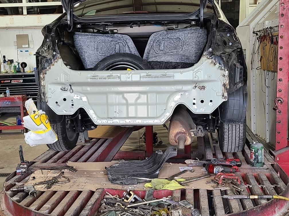 Rear view of a car with extensive damage in a repair shop. Trunk open, spare tire visible.