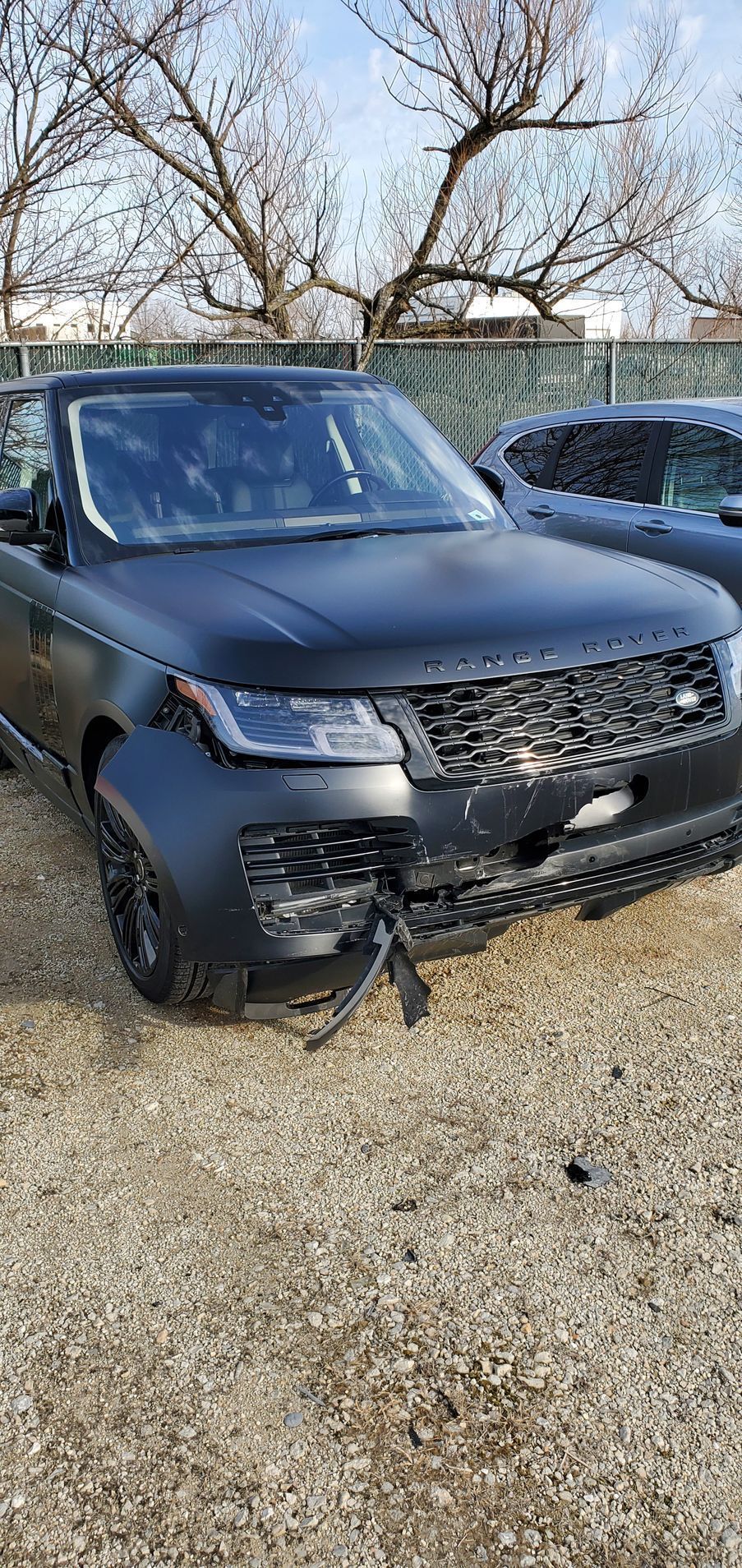 Black Range Rover with front-end damage parked outdoors. Gravel ground, bare trees in the background.