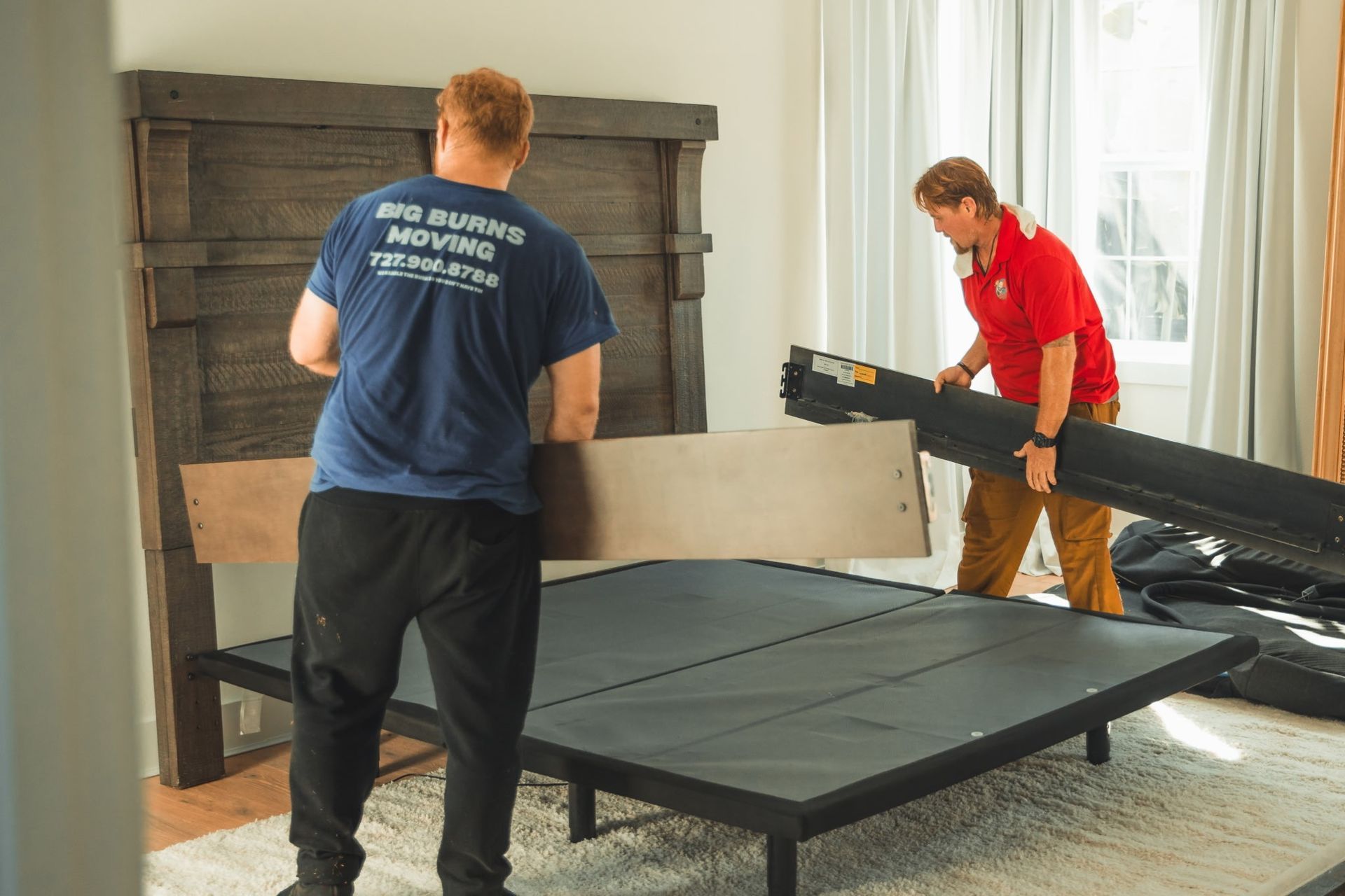 Two movers assembling a black bed frame in a bright bedroom.