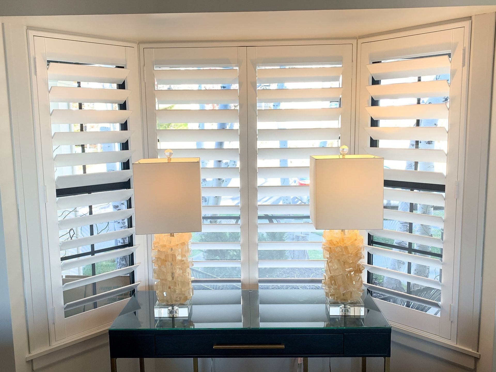 There are two lamps on a table in front of a window with shutters.