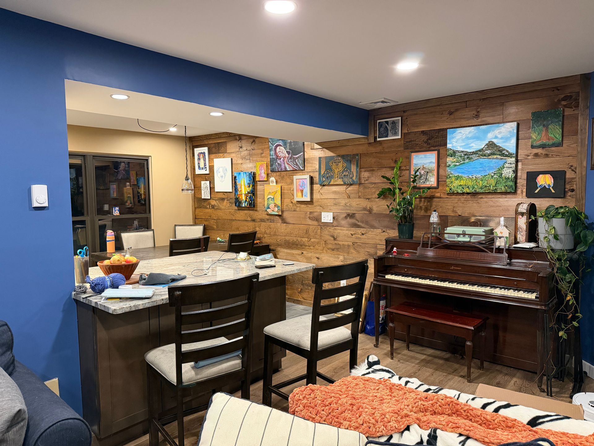 Kitchen area with blue walls, wood accent wall, paintings, piano, bar with stools.