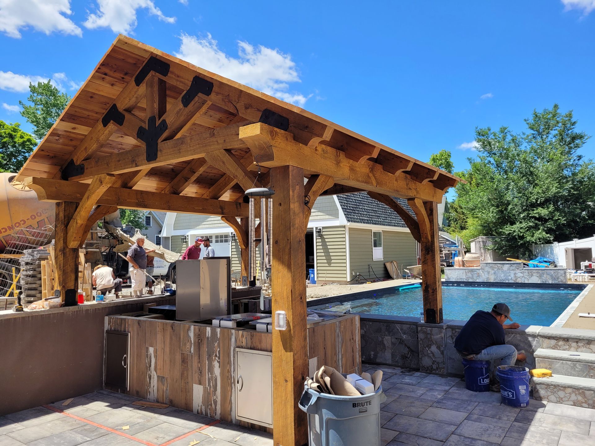 A man is sitting under a wooden structure next to a pool.