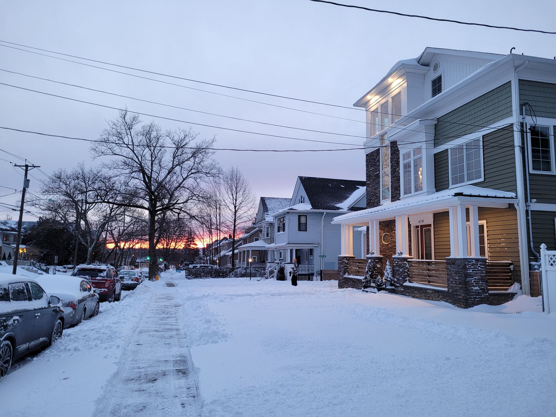 A snowy street with cars parked on the side of it and a sunset in the background.
