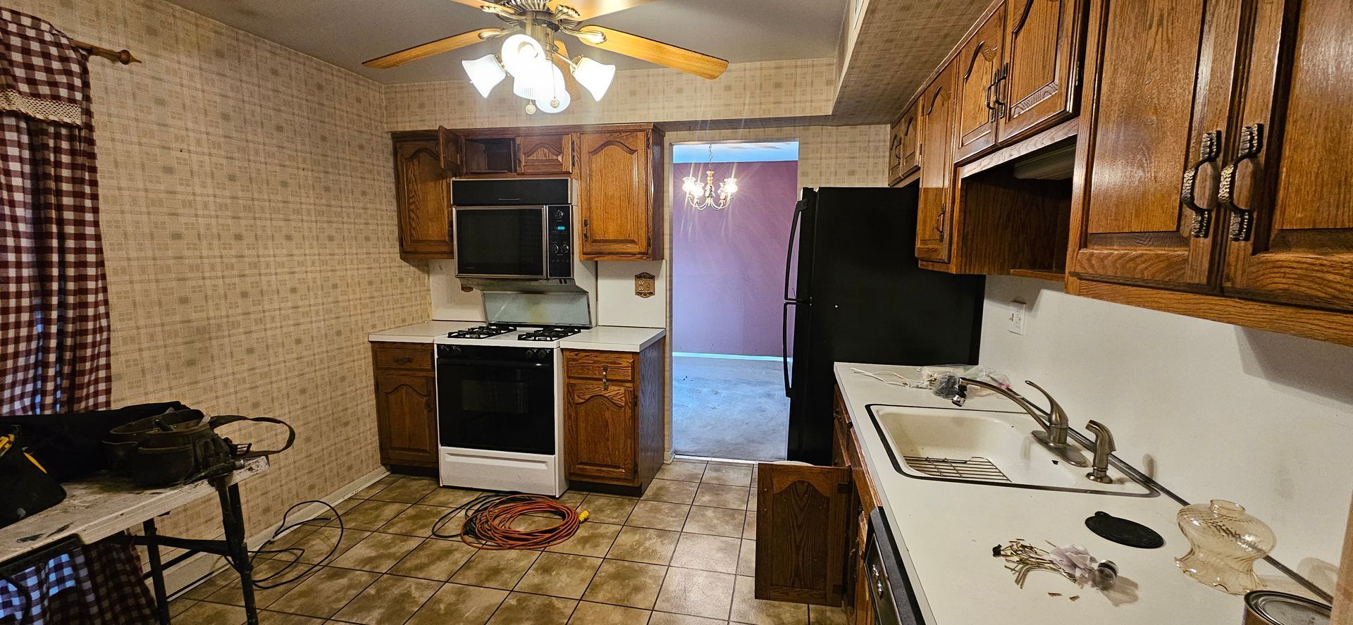 A kitchen with wooden cabinets, appliances, and patterned wallpaper. There is a doorway leading into a room.