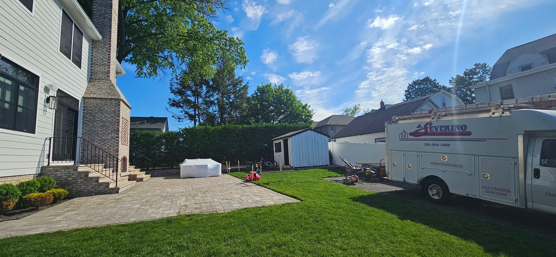 A backyard scene with a house, grass, trees, and a utility truck under a blue sky.