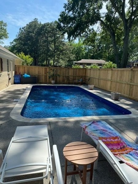 A rectangular pool with blue water surrounded by a concrete patio. Two white lounge chairs and a small table are in the foreground.