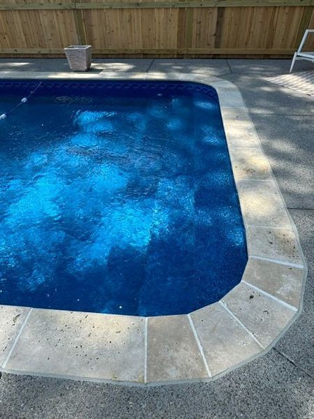 Blue swimming pool with concrete edging, partially covered with shadows, near a wooden fence.