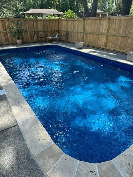 Rectangular swimming pool with blue water surrounded by concrete and a wooden fence.