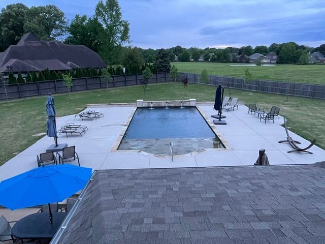Backyard with a rectangular swimming pool, concrete patio, lawn, and surrounding fence; cloudy sky.
