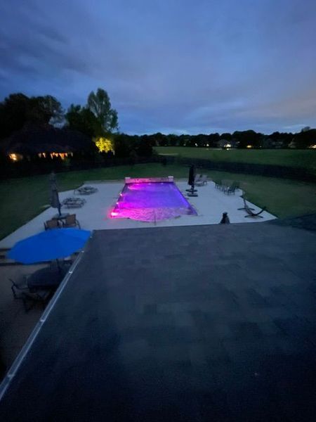 A rectangular pool illuminated with pink and purple lights at dusk. Trees and a house in the background.