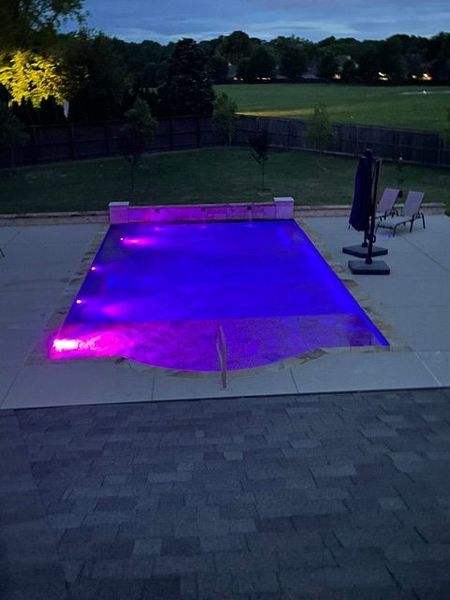Night view of a rectangular swimming pool with purple lights, surrounded by patio and green lawn.